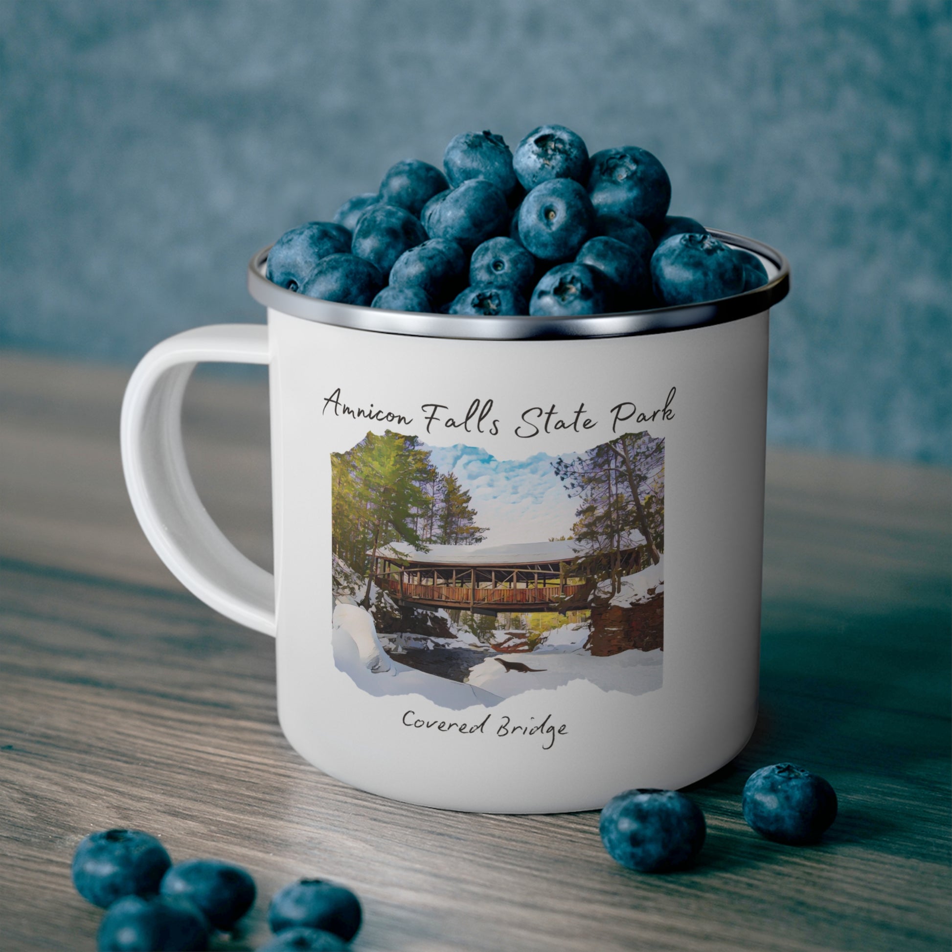 White enamel mug depicting the covered bridge at Amnicon Falls State Park in winter.  Background: mug sitting on a counter, full of blueberries.