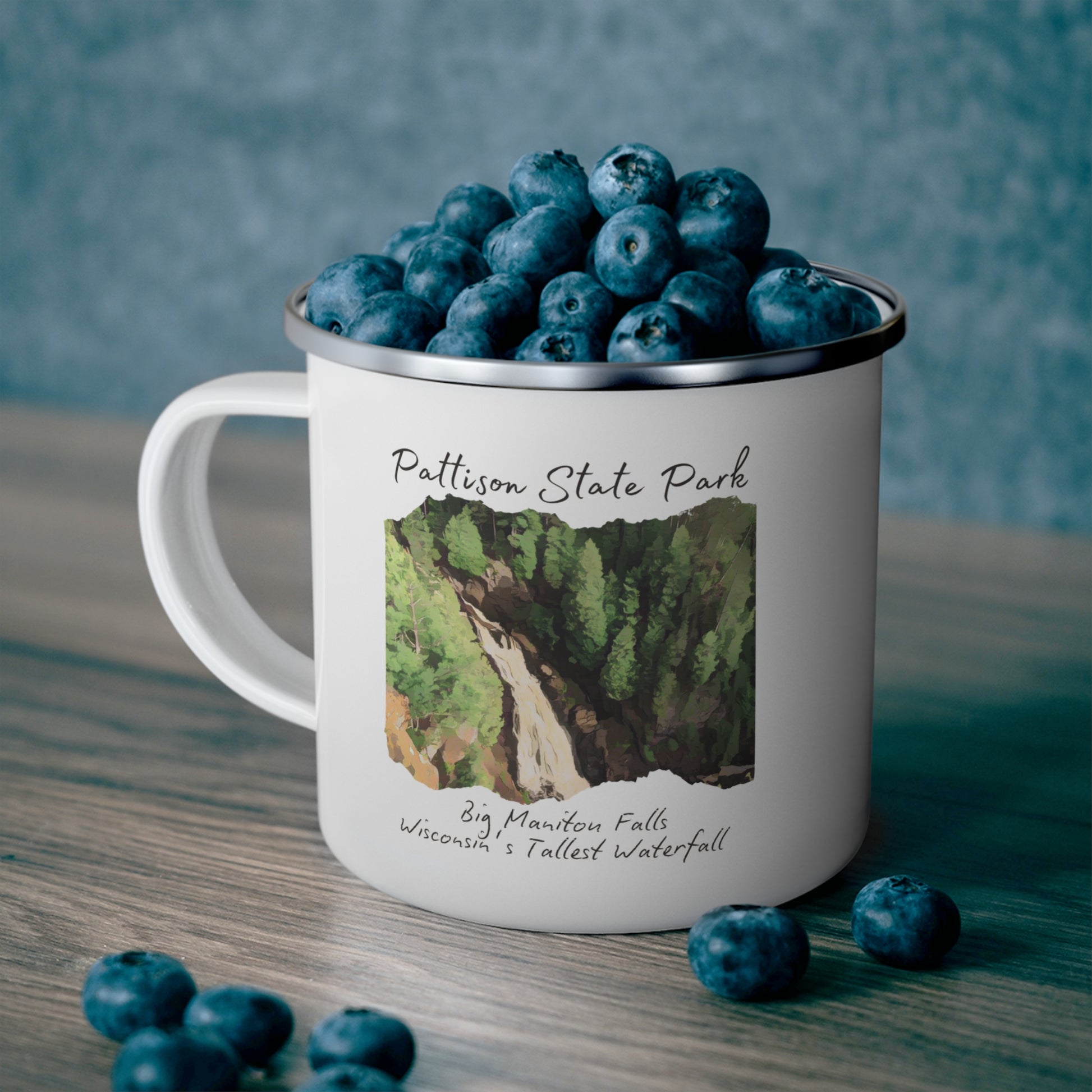 White enamel campfire mug with a CCC style rendering of Big Manitou Falls.  Mug contains the text: Pattison State Park, Big Manitou Fall, Wisconsin’s Tallest Waterfall.  Background of mug sitting on countertop, filled with blueberries.