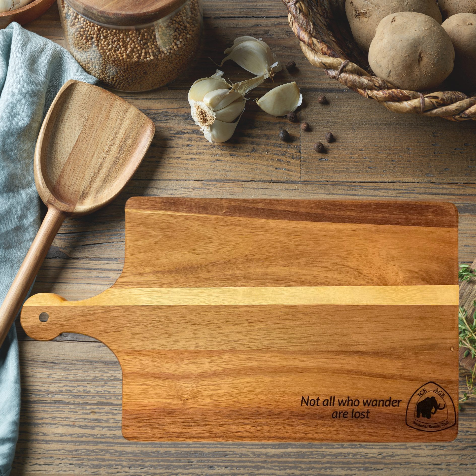 Acacia Wood cutting board with Ice Age Trail logo and text “Not all who wander are lost” engraved on the board.  Background of kitchen counter and utensils.