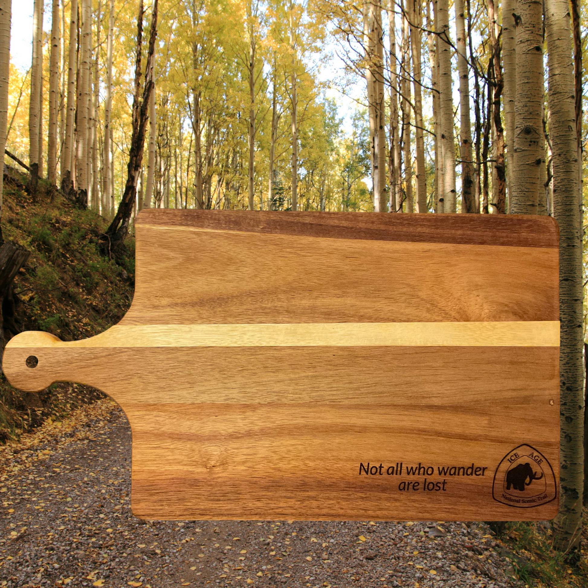 Acacia Wood cutting board with Ice Age Trail logo and text “Not all who wander are lost” engraved on the board.  Background of aspen trees.