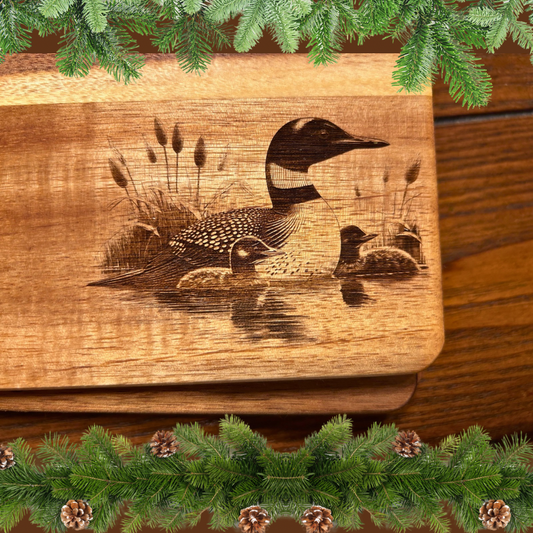 Acacia wood cutting board with a loon and two chicks engraved on it.  Evergreen garland background. 