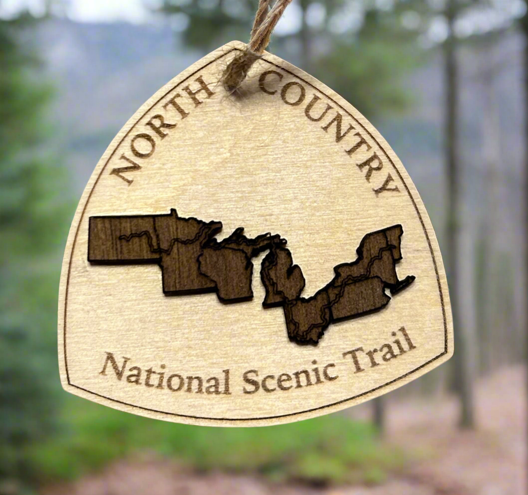 Wood ornament in the shape of the national trail's icon, with a walnut-colored North Dakota, Minnesota, Wisconsin, Michigan, Ohio, Pennsylvania, New York, and Vermont Shaped center piece depicting the North Country Trail Route.  Ornament contains the text "North Country National Scenic Trail". 