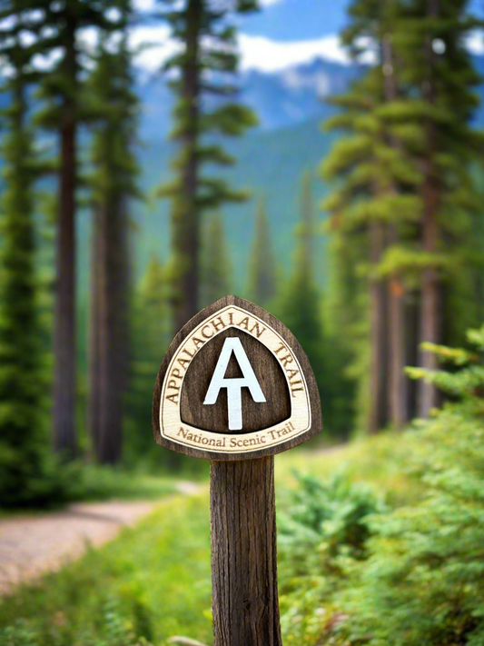 National scenic trail shaped magnet with Appalachian Trail National Scenic Trail, and white AT text symbol.  On a forest background with hiking trail.