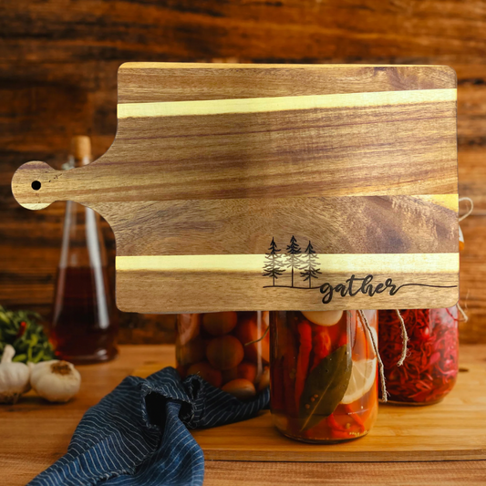 Acacia wood cutting board with three trees and the text gather engraved on the board.  Canned food and wine background. 