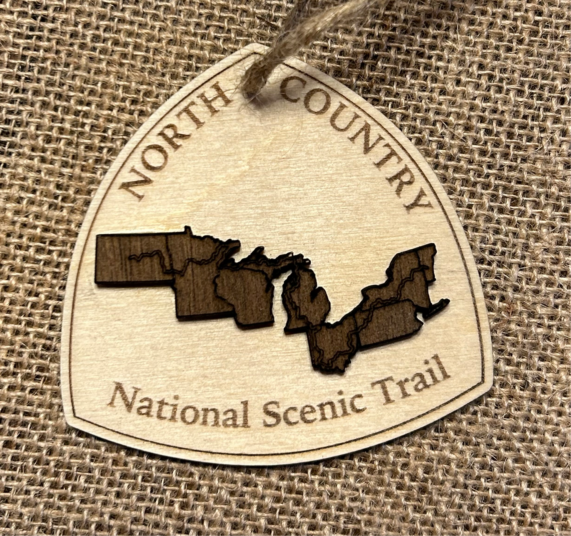 Wood ornament in the shape of the national trail's icon, with a walnut-colored North Dakota, Minnesota, Wisconsin, Michigan, Ohio, Pennsylvania, New York, and Vermont Shaped center piece depicting the North Country Trail Route.  Ornament contains the text "North Country National Scenic Trail". 