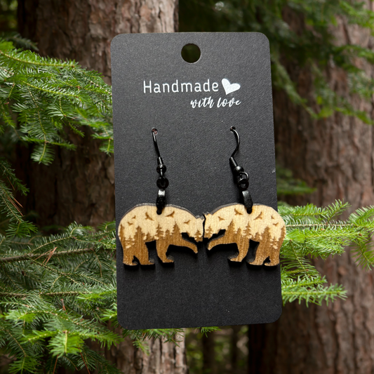 Bear shaped wood earrings with forest scene and flying birds engraved on them.  Photo background:  pine tree trunks and green needled branches.