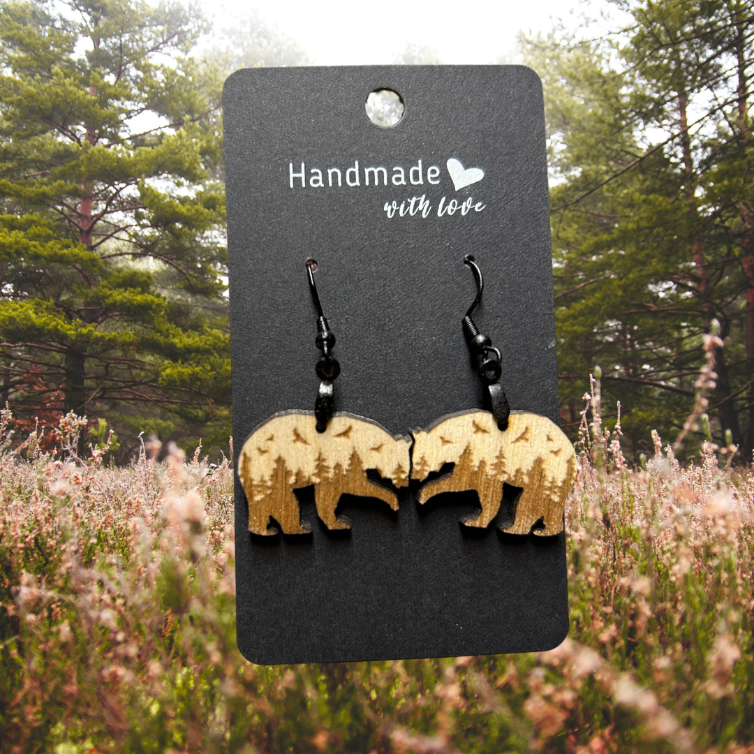 Bear shaped wood earrings with forest scene and flying birds engraved on them.  Photo background:  pine trees lining a misty meadow. 